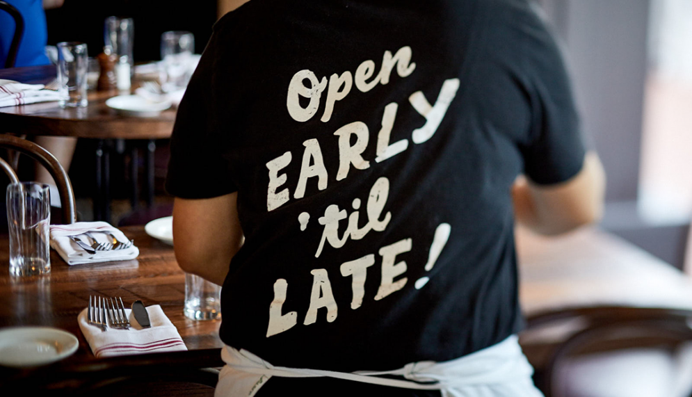 A photo of an employee setting a table with the back of the shirt reading " Open Early Til Late"
