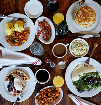 A photo of several brunch dishes, including eggs benedict, pancakes, waffles, omelet, home fries, bacon, coffee, and orange juice.
